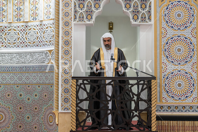 تعزيز الروحانية الجماعية, توجيه الناس نحو الحق والهداية, مفهوم العبادة والتقرب إلى الله تعالى, دور أئمة المساجد والخطباء في نشر الدين، إمام عربي خليجي إماراتي يرتدي البشت والكندورة والغترة البيضاء يقف أمام مكبر الصوت ويلقي الخطبة على المنبر