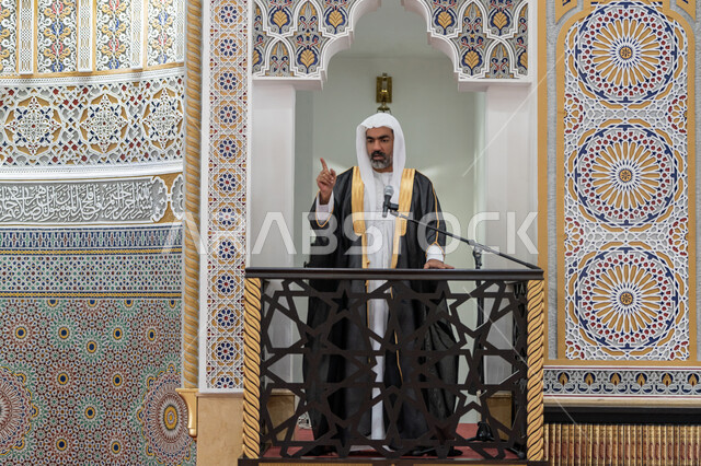 Calling Muslims to perform their duties, guiding people towards truth and guidance, the concept of worship and drawing closer to God Almighty, the role of mosque imams and preachers in spreading religion, an Arab Gulf Emirati imam wearing a bisht, kandura and white ghutra standing in front of a loudspeaker and delivering a sermon on the pulpit