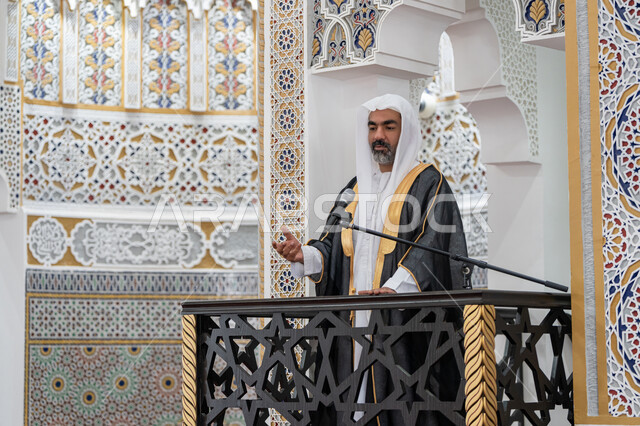 Calling Muslims to perform their duties, guiding people towards truth and guidance, the concept of worship and drawing closer to God Almighty, the role of mosque imams and preachers in spreading religion, an Arab Gulf Emirati imam wearing a bisht, kandura and white ghutra standing in front of a loudspeaker and delivering a sermon on the pulpit