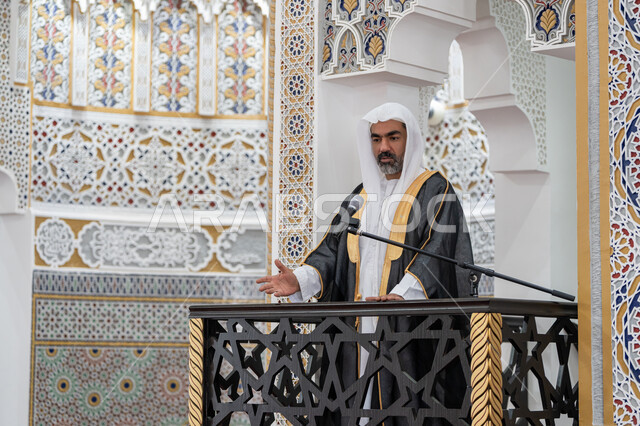 تعزيز الروحانية الجماعية, توجيه الناس نحو الحق والهداية, مفهوم العبادة والتقرب إلى الله تعالى, دور أئمة المساجد والخطباء في نشر الدين، إمام عربي خليجي إماراتي يرتدي البشت والكندورة والغترة البيضاء يقف أمام مكبر الصوت ويلقي الخطبة على المنبر