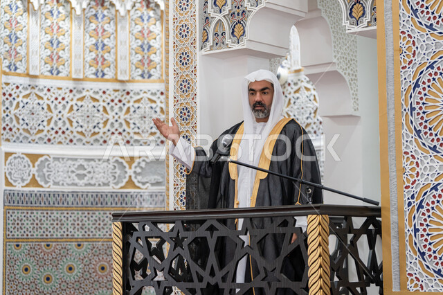 Strengthening collective spirituality, guiding people towards truth and guidance, the concept of worship and drawing closer to God Almighty, the role of mosque imams and preachers in spreading religion, an Arab Gulf Emirati imam wearing a bisht, kandura and white ghutra stands in front of a loudspeaker and delivers a sermon on the pulpit