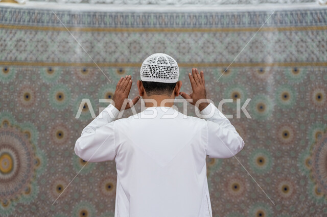 العبادة والتقرب إلى الله, إقامة الصلاة على وقتها، أداء الفرائض الدينية الإسلامية، الركن الثاني من أركان الإسلام، صورة مقربة من الخلف لرجل عربي خليجي إماراتي يرتدي الكندورة والقبعة البيضاء يقف باستقامة يرفع يديه لتكبيرة الإحرام, الشعور بالخشوع والطمأنينة