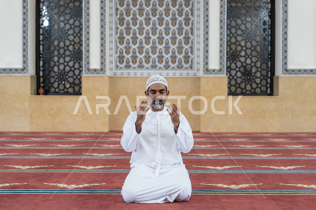 مفهوم الخشوع والتضرع, طلب الرحمة والمغفرة, العبادة والتقرب الى الله تعالى, رجل عربي خليجي اماراتي يرتدي الكندورة والقبعة البيضاء يجلس في إحدى المساجد ويرفع يديه للأعلى بتعابير الدعاء, أداء الفرائض الدينية الإسلامية، مفهوم أركان الإسلام