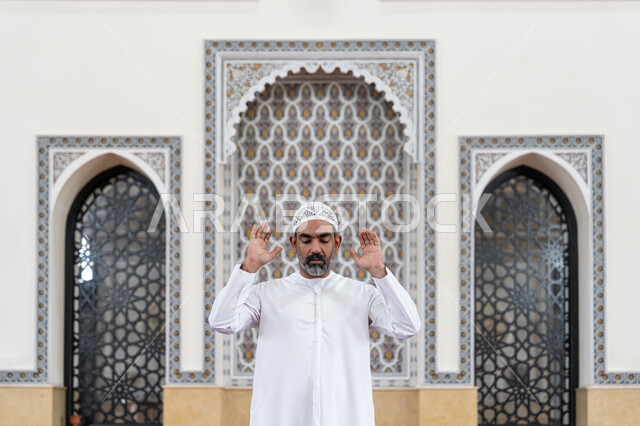 إقامة الصلاة على وقتها, العبادة والتقرب إلى الله، أداء الفرائض الدينية الإسلامية، الركن الثاني من أركان الإسلام، صورة مقربة لرجل عربي خليجي إماراتي يرتدي الكندورة والقبعة البيضاء يقف باستقامة يرفع يديه لتكبيرة الإحرام, الشعور بالخشوع والطمأنينة