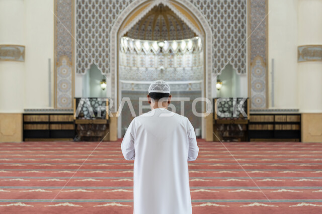 أداء الصلوات على وقتها, الشعور بالخشوع والطمأنينة, الركن الثاني من أركان الإسلام, العبادة والتقرب إلى الله تعالى, القيام بالفرائض الدينية الإسلامية، صورة من الخلف لرجل عربي خليجي إماراتي يرتدي الكندورة والقبعة البيضاء يقف باستقامة واعتدال لأداء فريضة الصلاة