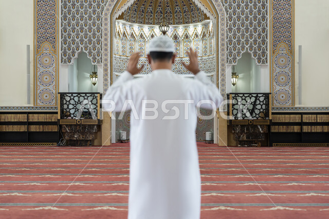 الركن الثاني من أركان الإسلام, إقامة الصلاة على وقتها, العبادة والتقرب إلى الله، أداء الفرائض الدينية الإسلامية، صورة من الخلف لرجل عربي خليجي إماراتي يرتدي الكندورة والقبعة البيضاء يقف باستقامة يرفع يديه لتكبيرة الإحرام, الشعور بالخشوع والطمأنينة