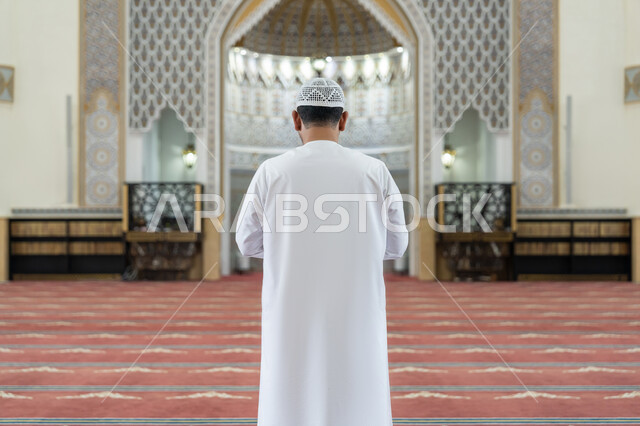 الشعور بالخشوع والطمأنينة, الركن الثاني من أركان الإسلام, العبادة والتقرب إلى الله تعالى, القيام بالفرائض الدينية الإسلامية، صورة من الخلف لرجل عربي خليجي إماراتي يرتدي الكندورة والقبعة البيضاء يقف باستقامة واعتدال لأداء فريضة الصلاة, أداء الصلوات على وقتها