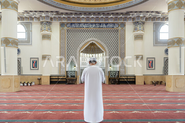القيام بالفرائض الدينية الإسلامية، الركن الثاني من أركان الإسلام, العبادة والتقرب إلى الله تعالى، صورة من الخلف لرجل عربي خليجي إماراتي يرتدي الكندورة والقبعة البيضاء يقف باستقامة واعتدال لأداء فريضة الصلاة, الشعور بالخشوع والطمأنينة, أداء الصلوات على وقتها