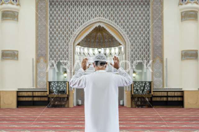 أداء الفرائض الدينية الإسلامية, إقامة الصلاة على وقتها, العبادة والتقرب إلى الله، الركن الثاني من أركان الإسلام، صورة من الخلف لرجل عربي خليجي إماراتي يرتدي الكندورة والقبعة البيضاء يقف باستقامة يرفع يديه لتكبيرة الإحرام, الشعور بالخشوع والطمأنينة