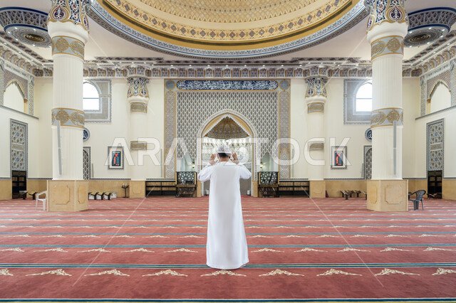 العبادة والتقرب إلى الله, إقامة الصلاة على وقتها، أداء الفرائض الدينية الإسلامية، الركن الثاني من أركان الإسلام، صورة من الخلف لرجل عربي خليجي إماراتي يرتدي الكندورة والقبعة البيضاء يقف باستقامة يرفع يديه لتكبيرة الإحرام, الشعور بالخشوع والطمأنينة
