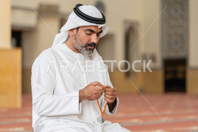 مفهوم التعبد في بيوت الرحمن, مفهوم التسبيح والتهليل, العبادة والتقرب الى الله تعالى, رجل عربي خليجي إماراتي يرتدي الكندورة والغترة البيضاء يجلس في إحدى المساجد ويمسك بيده سبحة، أداء الفرائض الدينية الإسلامية, أذكار بعد الصلاة