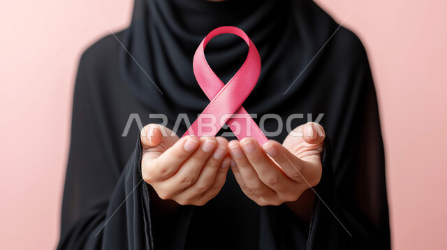 تحدي الأمراض الخبيثة، بورتريه مقرب ليدين إمرأة عربية خليجي سعودية ترتدي الحجاب والعباءة السوداء وتحمل الشريط الوردي، حملة توعوية للكشف المبكر عن سرطان الثدي، رمز الحملة العالمية، اليوم العالمي للتوعية والكشف المبكر، خلفية وردية