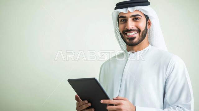 التسوق عبر الانترنت باستخدام المحمول، إنجاز المهام عن بعد، بورتريه مقرب لرجل عربي خليجي اماراتي يرتدي الكندورة والغترة البيضاء يحمل بيده جهاز التابلت، متابعة أخبار مواقع التواصل الاجتماعي، استخدام التكنولوجيا والتقنيات الحديثة، النظر الى الكاميرا بتعابير الفرح والسرور، خلفية بيضاء