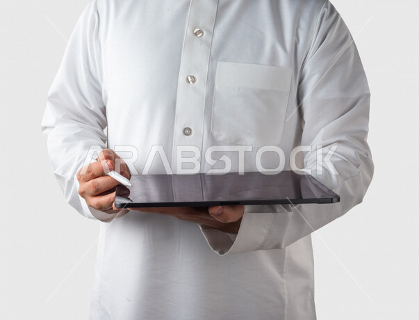 بورتريه لرجل خليجي سعودي يحمل بيده جهاز التابلت ، يستخدم القلم الخاص للضغط على شاشة الجهاز التقني ،تنظيم وادارة الاعمال عبر الجهاز التقني ، خلفية بيضاء