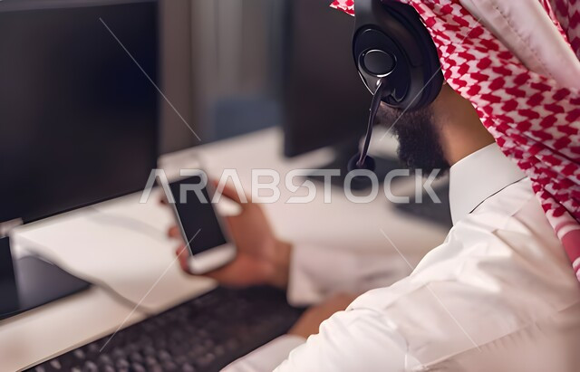 مفهوم خدمة العملاء، دمج العمل مع التقنية والتكنولوجيا, صورة مقربة من الخلف لموظف عربي خليجي سعودي يجلس على كرسي مريح يرتدي الشماغ والثوب التقليدي وسماعات الرأس في مركز الاستعلامات يقوم بالإجابة على الاسئلة والاستفسارات, اجراء اتصال فيديو من خلال الهاتف المحمول