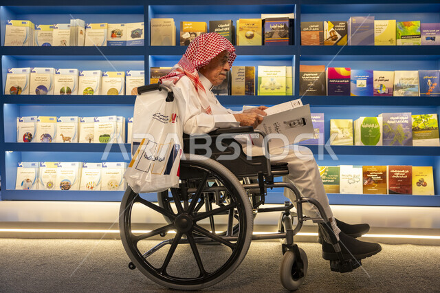 قضاء وقت الفراغ بالتعلم والثقافة، مفهوم محو الامية، هواية حب المطالعة والاستكشاف, صورة لرجل كبير السن مقعد عربي خليجي سعودي يرتدي الثوب التقليدي والشماغ يجلس على الكرسي يحمل بيده كتاب علمي تثقيفي في معرض الكتاب