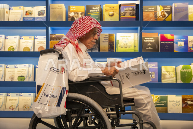 قضاء وقت الفراغ بالتعلم والثقافة، مفهوم محو الامية، هواية حب المطالعة والاستكشاف, صورة لرجل كبير السن مقعد عربي خليجي سعودي يرتدي الثوب التقليدي والشماغ يجلس على الكرسي يحمل بيده كتاب علمي تثقيفي في معرض الكتاب