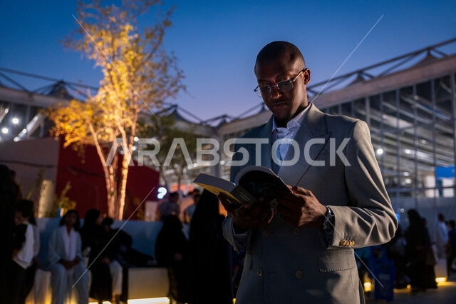 قضاء أوقات ممتعة في قراءة الكتب المفيدة للتعلم والتثقف، رجل عربي خليجي سعودي يرتدي الدلة الرسمية يقف ويحمل بيده كتاب، ممارسة هواية القراءة