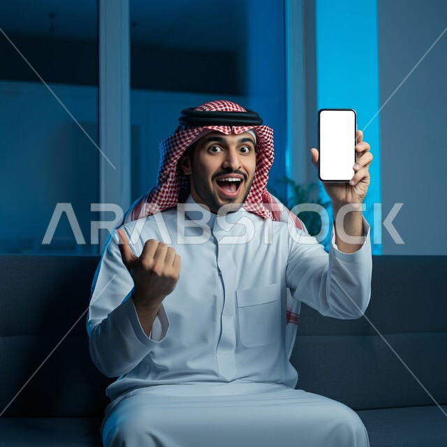 Admiration and satisfaction for achieving a certain thing, using advanced technology, a smiling young Saudi Arabian Gulf man wearing the traditional thobe and shemagh sitting on a comfortable sofa displaying a blank white screen via mobile phone raising his thumb up with gestures of surprise and astonishment