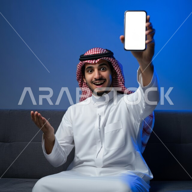 Looking at the camera with gestures of pleasure, using advanced technology for electronic digital marketing, displaying modern applications and services, a smiling young Saudi Arabian Gulf man wearing traditional thobe and shemagh displaying a blank white screen via mobile phone