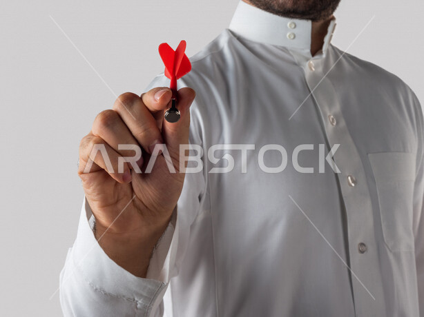 بورتريه لرجل خليجي سعودي يقوم بايماءات يد مختلفة ، يحمل بيده سهم مغناطيسي ، الدقة في تحديد الهدف ، المنافسة في العمل ، خلفية بيضاء