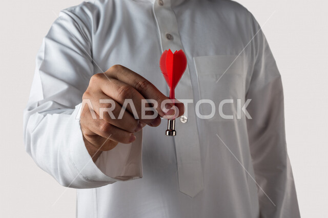 بورتريه لرجل خليجي سعودي يقوم بايماءات يد مختلفة ، يحمل بيده سهم مغناطيسي ، الدقة في تحديد الهدف ، المنافسة في العمل ، خلفية بيضاء