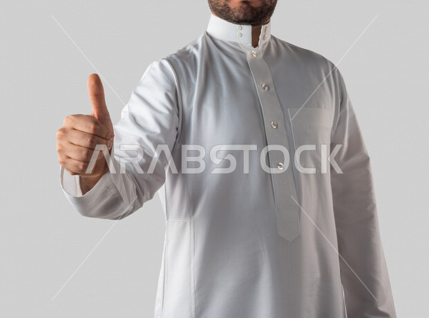 بورتريه لرجل خليجي سعودي يرتدي الزي الخليجي التقليدي يقوم بايماءات يد مختلفة ، يرفع اصبع الابهام لاعلى لاعجابه بشيء ما ، خلفية بيضاء