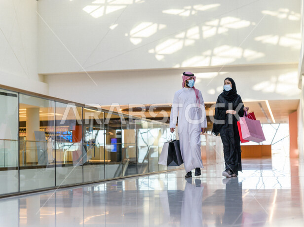 زوجان سعوديان يستمتعان بالتسوق معا في المركز التجاري , اتباع الاجراءات الاحترازية بلبس الكمامة , زوج وزوجته يحملان أكياس التسوق من المتجر , متعة التسوق في مولات السعودية .
