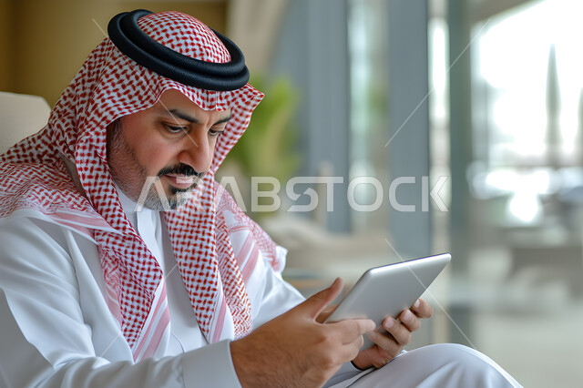 إجراء مقابلات عمل أونلاين, استخدام الأجهزة والتقنيات الحديثة لتسهيل الأعمال, صورة مقربة لرجل أعمال عربي سعودي خليجي يرتدي الثوب التقليدي والشماغ يحمل بيده جهاز التابليت، مهن ووظائف سعودية، ايماءات وتعابير التركيز والاندماج