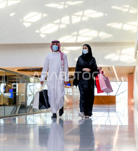 زوجان سعوديان يستمتعان بالتسوق معا في المركز التجاري , اتباع الاجراءات الاحترازية بلبس الكمامة , زوج وزوجته يحملان أكياس التسوق من المتجر , متعة التسوق في مولات السعودية .