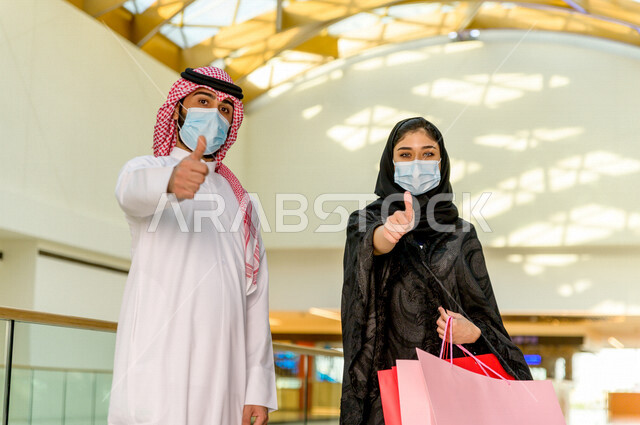 زوجان سعوديان يستمتعان بالتسوق معا في المركز التجاري , اتباع الاجراءات الاحترازية بلبس الكمامة , زوج وزوجته يحملان أكياس التسوق من المتجر , متعة التسوق في مولات السعودية .