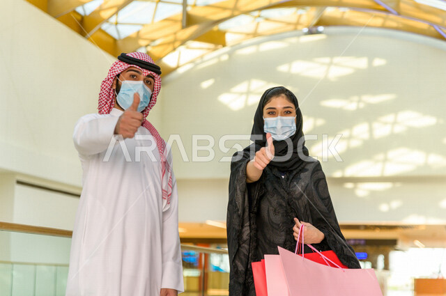 زوجان سعوديان يستمتعان بالتسوق معا في المركز التجاري , اتباع الاجراءات الاحترازية بلبس الكمامة , زوج وزوجته يحملان أكياس التسوق من المتجر , متعة التسوق في مولات السعودية .