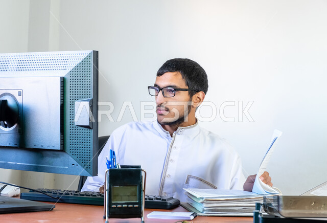 مهن ووظائف إدارية مكتبية، ايماءات الاندماج والتركيز، دمج العمل مع التقنية والتكنولوجيا الحديثة، شاب عربي خليجي سعودي يرتدي الثوب التقليدي يجلس خلف المكتب ويعمل على جهاز الحاسوب، مراجعة التقارير والملاحظات من الملفات، مفهوم ادارة الاعمال وتحقيق اهداف الشركة