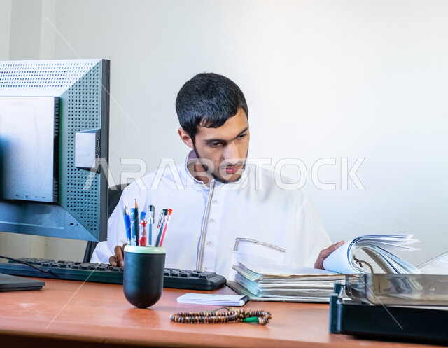 Integrating work with technology and modern technology, a young Saudi Arabian Gulf man wearing traditional clothing sits behind a desk and reviews reports and notes from files, the concept of business management and achieving company goals, office administrative jobs and professions, gestures of integration and focus