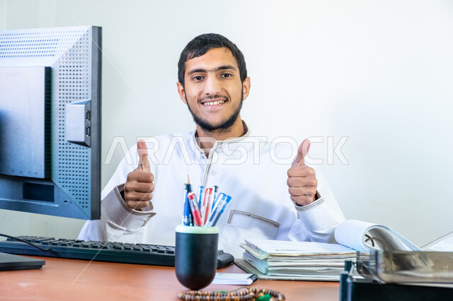 Professional computer and information technology jobs, a young Saudi Arabian Gulf man wearing traditional thobe sitting behind a desk and raising his thumbs in gestures of quality and excellence, browsing the internet and working on a laptop, office administrative jobs and professions, the concept of business administration and achieving company goals