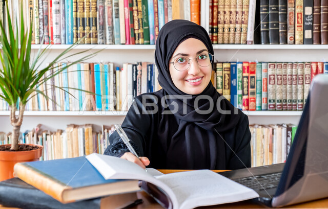 The concept of culture and self-learning, a close-up of a smiling veiled Saudi Arabian Gulf young woman wearing a black abaya looking at the camera with gestures of happiness, using modern technical devices to follow university lectures, the concept of distance education, curricula and educational materials in Saudi Arabia