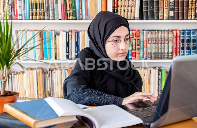 المحاضرات الجامعية الإلكترونية والتعلم عن بعد، التعليم العالي في الكليات السعودية، مفهوم الثقافة والتعلم الذاتي، كتابة التقارير والأبحاث العلمية، شابة عربية خليجية سعودية تجلس في مكتبة الجامعة وتستخدم الحاسوب المحمول في مراجعة الدروس اليومية