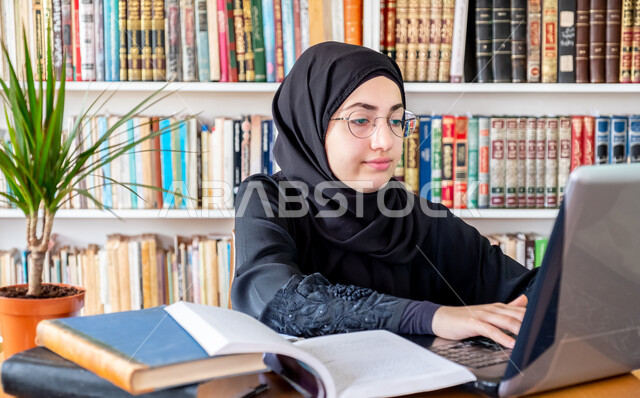 مراجعة الدروس اليومية باستخدام الحاسوب المحمول، المحاضرات الجامعية الإلكترونية والتعلم عن بعد، التعليم العالي في الكليات السعودية، مفهوم الثقافة والتعلم الذاتي، شابة عربية خليجية سعودية تجلس في مكتبة الجامعة تقوم بكتابة التقارير والأبحاث العلمية