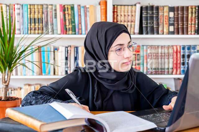 التعليم العالي في الكليات السعودية، مفهوم الثقافة والتعلم الذاتي، كتابة التقارير والأبحاث العلمية، شابة عربية خليجية سعودية تجلس في مكتبة الجامعة وتستخدم الحاسوب المحمول في مراجعة الدروس اليومية، المحاضرات الجامعية الإلكترونية والتعلم عن بعد