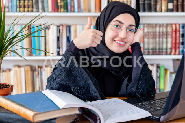 التعليم عن بعد، المناهج والمواد التعليمية في السعودية، مفهوم الثقافة والتعلم الذاتي , صورة مقربة لشابة عربية خليجية سعودية محجبة مبتسمة ترتدي عباءة سوداء ترفع اصبع الابهام بعلامة الامتياز والرضا، استخدام الأجهزة التقنية الحديثة لمتابعة المحاضرات الجامعية