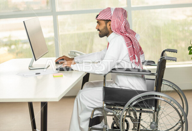 موظف عربي سعودي خليجي من ذوي الهمم يعمل على جهاز الحاسوب في مقر العمل, دمج و توظيف ذوي الاعاقه في مختلف المجالات, التمكين الاجتماعي, اهتمام  ودعم المجتمع السعودي لذوي الاحتياجات الخاصه, شعور ذوي الهمم بالراحه والطمأنينه في بيئتهم
