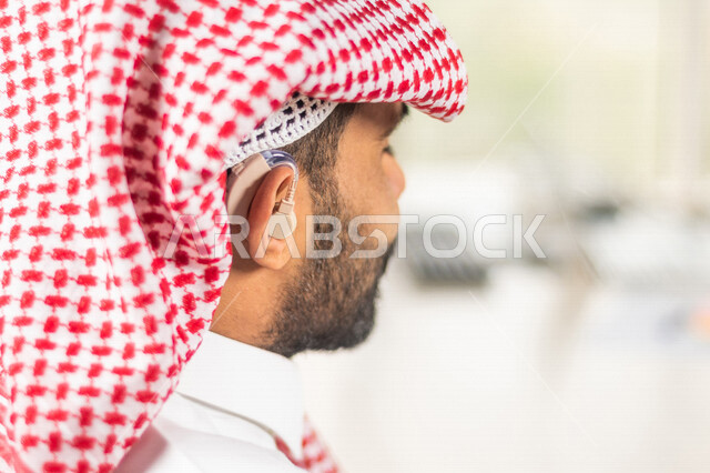 موظف عربي سعودي خليجي مصاب بالصمم يرتدي سماعة الاذن لتساعده على السمع, رجل عربي سعودي خليجي من ذوي الهمم في مقر عمله, دمج ذوي الاعاقه في مختلف المجالات, مساعدات الصم بالسماعه الطبيه