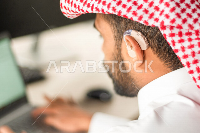 موظف عربي سعودي خليجي مصاب بالصمم يرتدي سماعة الاذن لتساعده على السمع, رجل عربي سعودي خليجي من ذوي الهمم في مقر عمله, دمج ذوي الاعاقه في مختلف المجالات, مساعدات الصم بالسماعه الطبيه