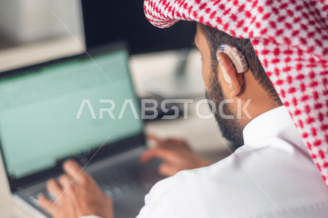 موظف عربي سعودي خليجي مصاب بالصمم يرتدي سماعة الاذن لتساعده على السمع, رجل عربي سعودي خليجي من ذوي الهمم في مقر عمله, دمج ذوي الاعاقه في مختلف المجالات, مساعدات الصم بالسماعه الطبيه