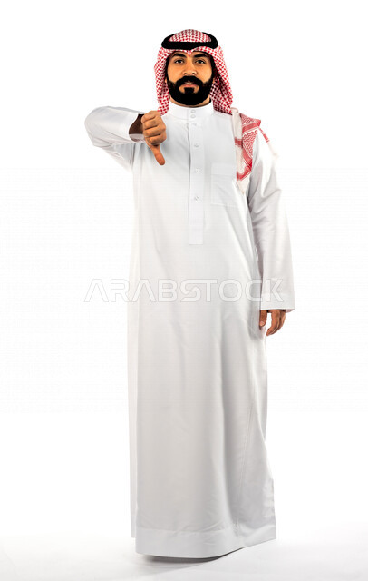 بورتريه لرجل سعودي خليجي يشير بإصبعه الإبهام لأسفل بإيماءات وجه ويدين مختلفة تدل على الرفض وعدم الموافقة ، خلفية بيضاء لرجل سعودي مسلم يقوم بجلسة تصوير ، يرتدي الزي التقليدي السعودي مع شماغ وعقال