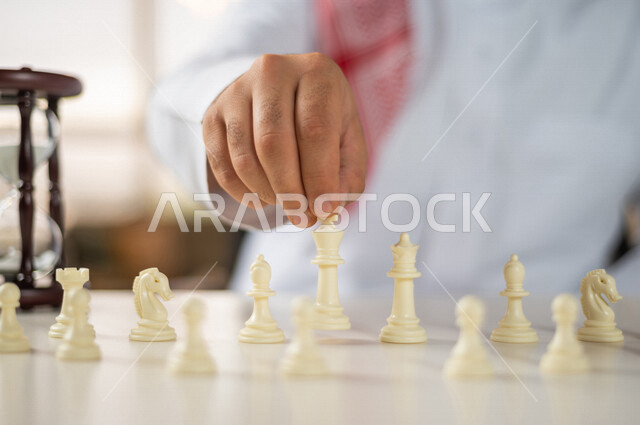 صورة مقربة ليد رجل سعودي خليجي يلعب لعبة الشطرنج، مفهوم التركيز والتحدي، التسليه والترفيه، لعبة شطرنج على طاولة بيضاء، مفهوم التخطيط والتفكير في استراتيجية مناسبة، ساعة رملية  خشبية (البنكام) على الطاولة، مفهوم إدارة الأعمال والوقت