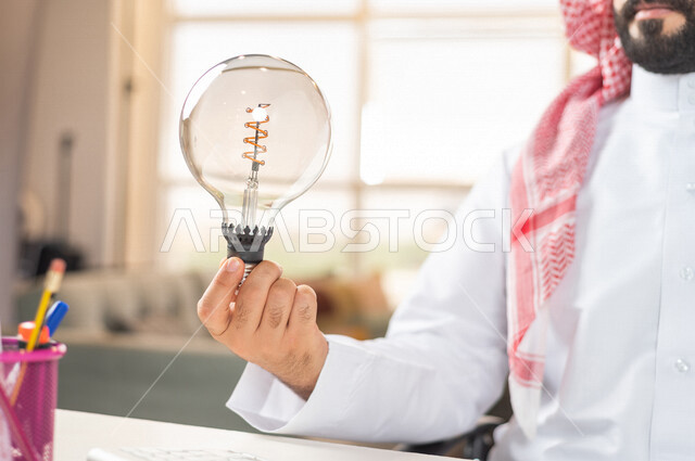 صورة مقربة ليد رجل سعودي خليجي يحمل المصباح الضوئي ، أفكار إبداعية تطويرية ، مفهوم الفكرة مع الإبتكار والإلهام ، تحقيق الإنجازات والإبتكارات ، براءة الإختراع ، المصباح الكهربائي الضوئي