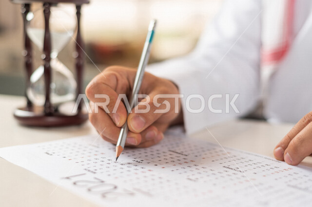 صورة مقربة ليد رجل سعودي خليجي يستخدم القلم لجدول الكتابة في التقويم، الساعة الرملية الزجاجية (البنكام) على الطاولة ، مفهوم إدارة الأعمال والوقت، توقيت الرمل، تحديد مواعيد العمل ، مفهوم إدارة الجدول الزمني ، قياس الوقت