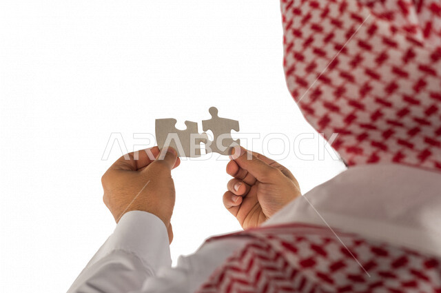 بورتريه بصورة مقربة ليد رجل سعودي خليجي يحمل قطعتين من اللغز يحاول ربط وتوصيل قطع اللغز الصغيرة ، مفهوم التركيز وحل اللغز ، التسليه والترفيه ، لعبة بازل ، لعبة تركيب الصور على خلفية بيضاء