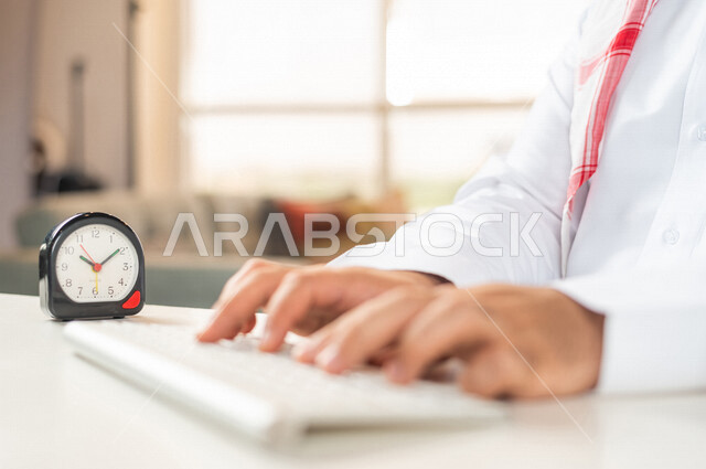 صورة مقربة لرجل أعمال سعودي خليجي ، استخدام لوحة المفاتيح للكتابة ، ساعة رقمية صغيرة على الطاولة ، مفهوم إدارة وقت العمل ، الدقة في المواعيد ، التذكيرات والتنبيهات ، العمل عن بعد ، التسويق الإلكتروني ، إنجاز المهامات في الوقت